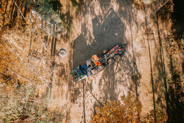 Obraz premium Aerial shot showing a truck with logging machinery on a trailer in a forest clearing, surrounded by trees and shadows on the ground.