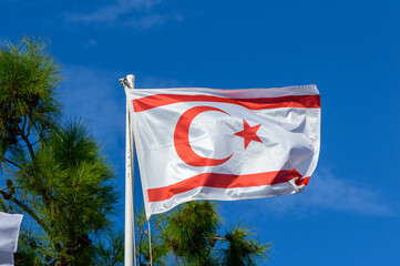 The vibrant flag of Northern Cyprus waving proudly against a brilliant blue sky, surrounded by lush greenery on a sunny day