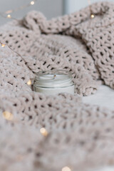 A candle in a glass jar with a wooden wick stands among a knitted plush soft warm plaid. Warm comfort. Vertical photo