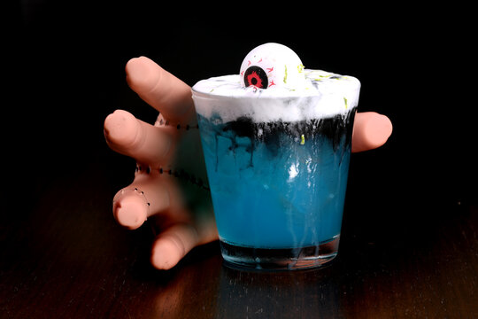 hand picking up drink blue cura&ccedil;ao cocktail with decorative eye and ginger spuma at halloween party with blurred background