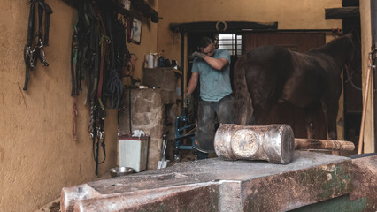A Farrier with a Forging Hammer Shoeing a Horse in a Stable