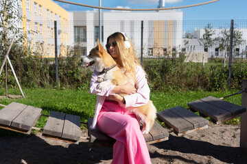 Happy young woman girl in sunglasses and headphones is petting her cute corgi dog puppy. Best friends. Female pet owner and her dog enjoying the sunny weather.