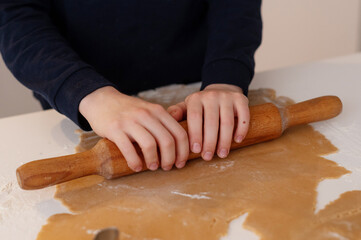 A handsome dark-haired boy with beautiful eyes and a smile in New Year's pajamas, rolls out gingerbread dough in the kitchen with a rolling pin, a Christmas tree stands in the background and a garland