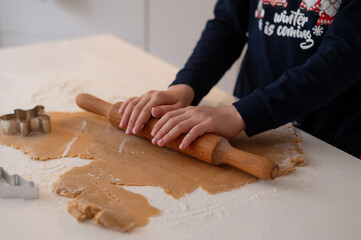 A handsome dark-haired boy with beautiful eyes and a smile in New Year's pajamas, rolls out gingerbread dough in the kitchen with a rolling pin, a Christmas tree stands in the background and a garland