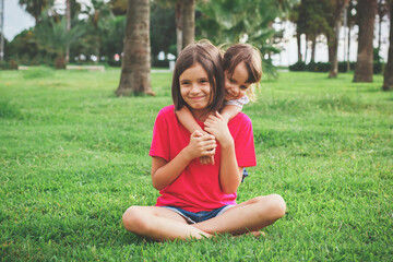 two girls 7 and 2 years old, two sisters sitting on green grass in a park on a summer day, the younger one hugging the older one, sibling relationship concept