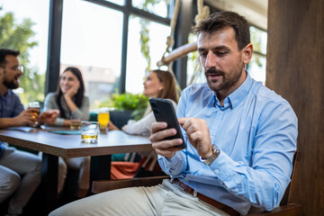 Young businessman using his phone while in restaurant with his friends in the background.