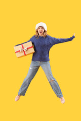 Jumping young woman in Santa hat with Christmas gift on yellow background
