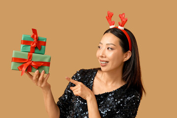 Young Asian woman in reindeer horns with Christmas gift boxes on brown background