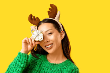 Happy young Asian woman in reindeer horns with Christmas gift box on yellow background