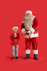 Santa Claus and cute little boy with milk and cookies on red background