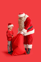 Santa Claus and cute little boy with bag full of Christmas gifts on red background