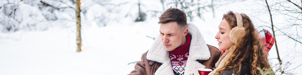 Fototapeta premium Young loving couple spending winter holidays in countryside cottage house. Wearing warm clothes, knitted sweater, mittens. Hot drinks. Cozy Christmas atmosphere, New Year's Eve snowy frosty day banner