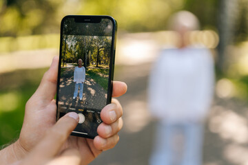 Closeup of unrecognizable senior woman posing happily in sunny park, while husband photographer capturing moment with smartphone, lush green background adding to serene scene, selective focus.