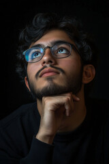 Obraz premium A young Hispanic man with glasses and a beard gazes into the distance with a thoughtful expression, his chin resting on his hand. The dark background enhances the quiet, contemplative atmosphere.