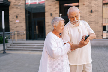 Portrait of beautiful elderly couple happily exploring new city streets, relying on smartphone for directions. Gray-haired senior male and female navigating unfamiliar city with smartphone navigation.