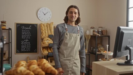 Young man with long hair wearing an apron making a beckoning gesture with his hand in a bakery full of breads and pastries.