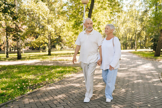 Full length portrait of senior couple enjoying leisurely stroll in green park, holding hands intertwined, sharing tender moment on sunny day. Active elderly man and woman enjoying retirement together.