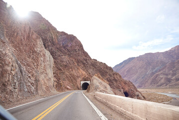 Road trip scenery in Mendoza, Argentina: an empty road in the middle of a mountainous landscape, on the right the river and in front a tunnel through the mountain.