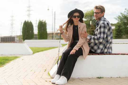 A visually impaired woman and her partner sit together outside, engaging in a warm conversation. They enjoy each other's company, surrounded by a tranquil landscape under a bright sky