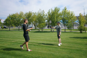 Fototapeta premium In a lush park, two friends play badminton on a beautiful sunny day. The vibrant greenery and clear sky create a perfect backdrop for their friendly match