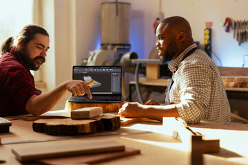 Cabinetmaker and colleague using CAD software on laptop to design wooden objects. Master carpenter and apprentice using program on notebook to plan furniture assembling in joinery