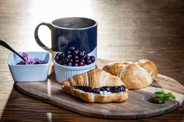 A plate of croissants with huckleberry jam and a cup of coffee sit on a wooden table. The croissants are arranged on a wooden tray, and the coffee is in a blue mug. Concept of comfort and relaxation