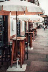 Outdoor cafe with wooden high tables, bar stools, and white umbrellas lining a city street. Shallow depth of field emphasizes the distant street. Selective focus on front table