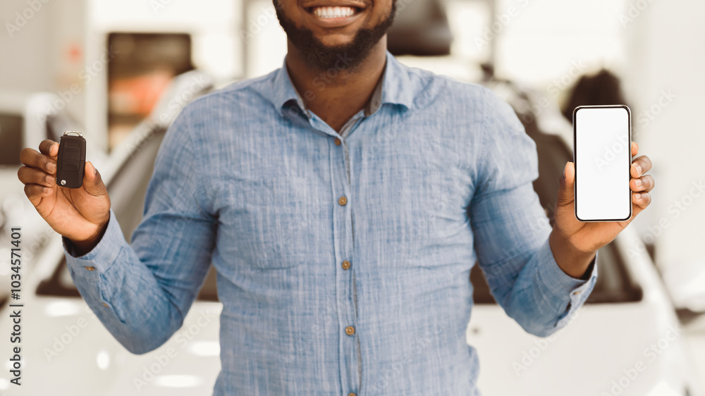 Poster unrecognizable black man showing new car key and smartphone with blank screen standing in automobile