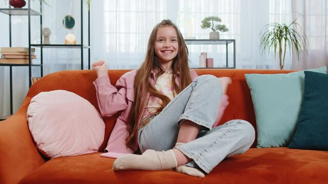 Happy girl using mobile smartphone typing playing browsing found out great big surprise, good positive news, celebrate online game win. Female teenager kid child clapping in living room sits on sofa.