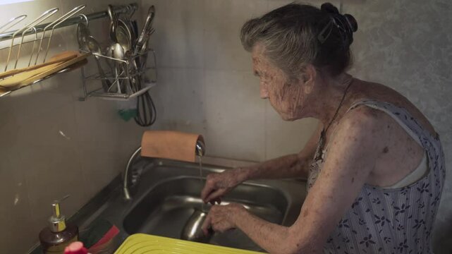 Poor senior woman with sad emotions, gray hair and deep wrinkles, 90 years old, washes and wipes dishes in old kitchen. Sad elderly female cleans crockery lonely in small kitchen. Poverty and misery.