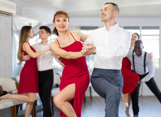 Fototapeta premium Cheerful woman with bright makeup, in stylish red dress, dancing playful lindy hop paired with male partner during group training in dance studio..