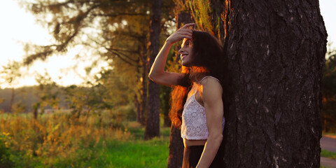 Beautiful happy smiling woman standing the back to the oak tree and looking on amazing summer sunset in the orange sun rays on the field with trees and green grass. Atmospheric summer