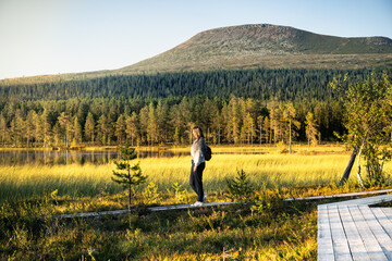 Woman walking on a wooden path beside a serene lake in the Idre wetlands, with the majestic Mount Städjan rising in the background during the golden hour of summer in Dalarna Sweden