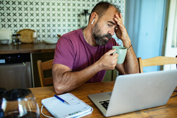 Man stressed while working on laptop with coffee in hand at kitchen