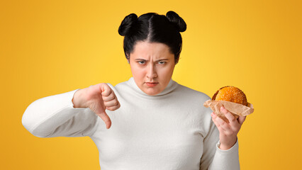 Stop Eating Fast Food. Annoyed overweight girl holding hamburger and showing thumb down, rejecting unhealthy meal, yellow background with free space