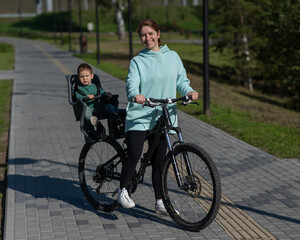 Obraz premium Caucasian woman riding bicycle with toddler son sitting behind her in child seat in park. 