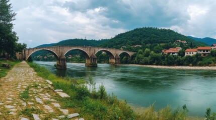 Fototapeta premium A scenic view of a stone bridge over a river surrounded by green hills and houses.