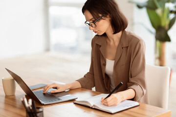Female Career Concept. Beautiful young woman in specs sitting at wooden desk with laptop and writing in notebook