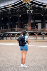 Unrecognizable young Latina woman in casual summer clothes, holding a smartphone, explores the historic Higashi Hongan-ji Temple in Kyoto, Japan, with a backpack and a cheerful expression