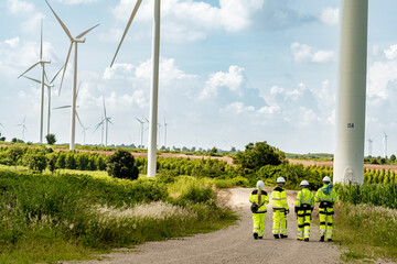  group of engineers in high visibility gear stands at a wind farm, observing several wind turbines....