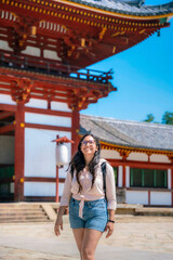 A happy young Latina woman, dressed casually with glasses and a backpack, smiles while exploring the historic Todaiji Kondo Hakkaku Toro in Nara, Japan, on a sunny day.