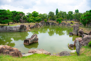 A peaceful view of the tranquil pond surrounded by rocks and greenery in the gardens of Nijo Castle, Kyoto, Japan. The calm water reflects the lush trees and serene atmosphere.