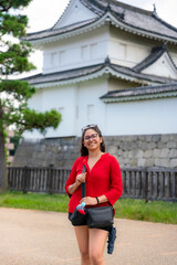 A smiling Latina tourist stands in front of the historic Nijo Castle in Kyoto, Japan. Dressed in a...