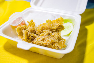 close up, single fried chicken with cucumber slices and raw chili sauce on a white styrofoam plate, with a yellow cloth background. Focus on fried chicken. noisy, similar others, exposure