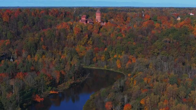 Aerial view of Turaida Castle's red brick towers amidst vibrant autumn foliage in Sigulda, Latvia, with Gauja National Park in the background.