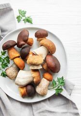 A variety of fresh mushrooms displayed on a plate surrounded by green herbs on a simple wooden surface