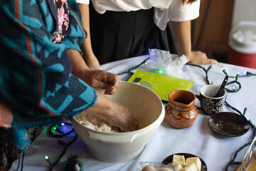 Hands of an unrecognizable older woman, with a young woman, making a dough by mixing it in a bowl, preparing it with other ingredients and utensils in a rustic kitchen on a festive table.