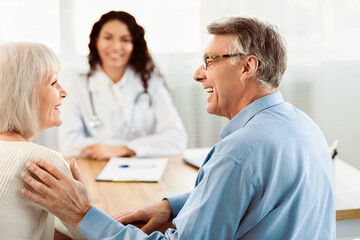 Fototapeta premium Good Diagnosis. Happy elderly couple looking at each other during consultation with female doctor at clinic, free space
