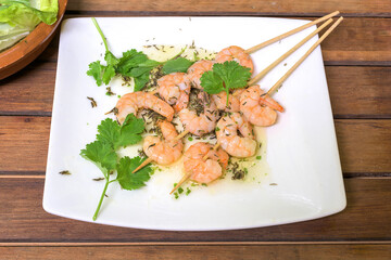 Garlic prawn skewer with olive oil, cilantro and lettuce on a white plate on a wooden background