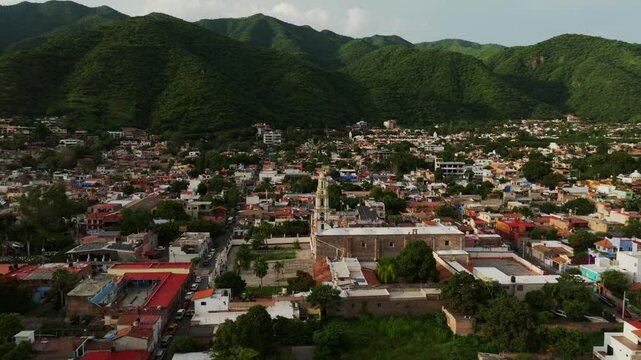 Panoramic aerial orbit of San Andres Apostol facade and lake Chapala, with mountains on the horizon and lush green vegetation in rainy season